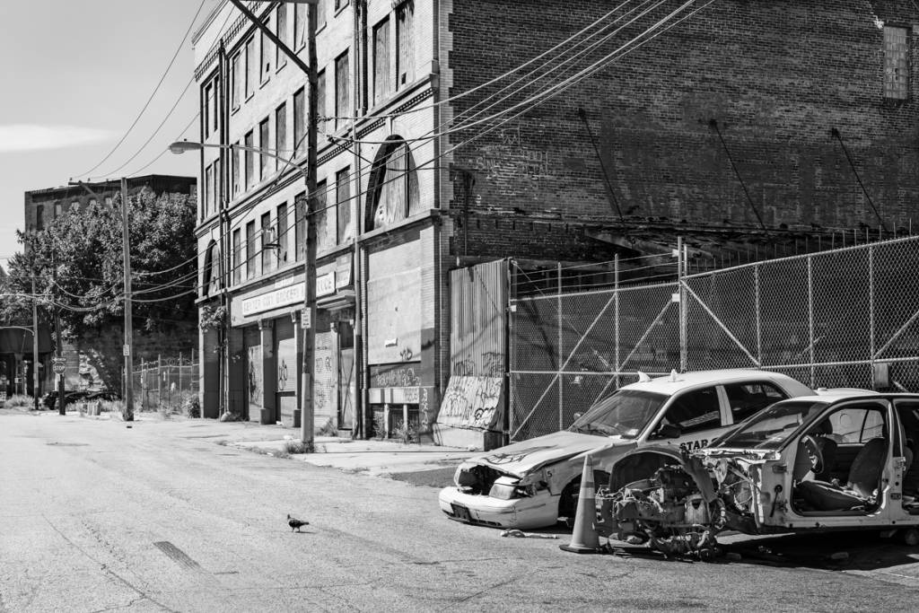 Black and white view of stripped car bodies on street next to shabby building.