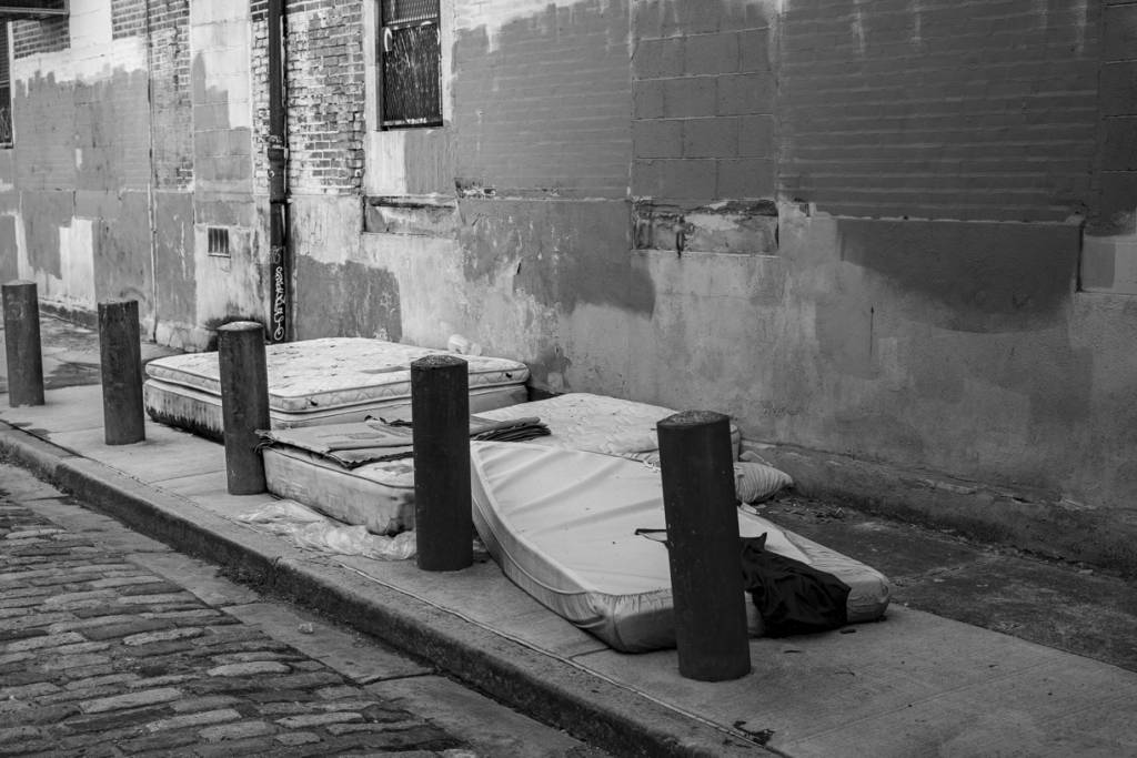 Black and white view of mattresses on back street sidewalk where homeless sleep.