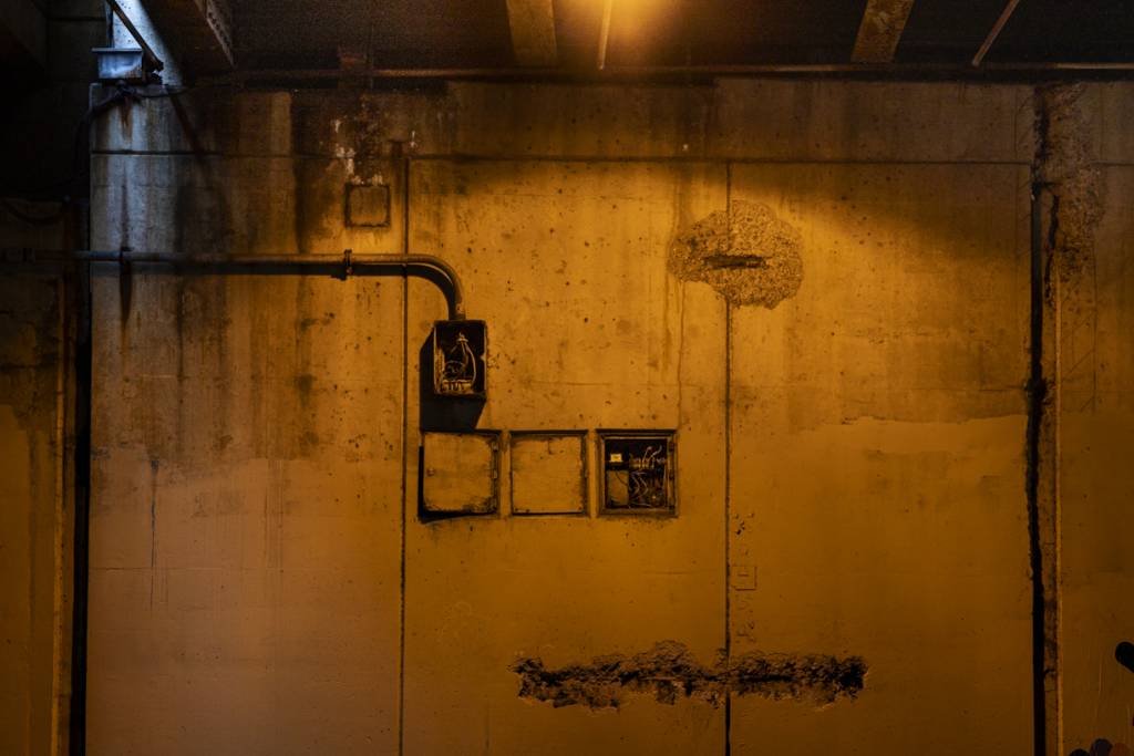 Color view of concrete wall below overpass showing electrical connection boxes and blue light in corner.